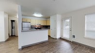This single-floor space offers a practical and well-designed open-concept layout that seamlessly connects the kitchen and living area. The kitchen is equipped with stainless steel appliances, including a refrigerator and an electric range with an overhead ventilation hood. Light-toned cabinetry lines the walls, providing ample storage, while a mosaic tile backsplash adds a subtle decorative touch. A peninsula with a gray countertop extends into the living space, creating a natural breakfast bar that enhances both functionality and flow between areas. Wood-style laminate flooring runs throughout the space, lending warmth and cohesion to the interior. A white paneled door near the kitchen leads to what appears to be additional storage. Natural light filters into the space through a windowed exterior door fitted with blinds, brightening the area and offering a connection to the outdoors. The walls are finished in a clean, neutral white, contributing to an overall bright and airy ambiance. Flush-mount ceiling lighting ensures consistent illumination throughout. The space presents a clean, move-in-ready environment suited for everyday living.