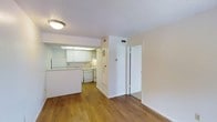 This single-floor space showcases a practical and well-organized open-concept layout, where the living area flows seamlessly into the kitchen. The kitchen is equipped with white cabinetry, a white refrigerator, a dishwasher, and a range, all complemented by light-toned countertops. A pass-through counter connects the kitchen to the adjacent living space, enhancing the open feel of the interior. Overhead, a dropped ceiling with panel lighting brightens the kitchen workspace effectively.

The living area features warm hardwood flooring that extends throughout the unit, adding a natural, inviting tone to the overall aesthetic. Walls are finished in a clean, neutral white, contributing to a bright and airy atmosphere. A textured ceiling adds subtle visual interest to the space. Two louvered doors are visible—likely leading to a closet and storage areas—providing functional storage solutions. A standard flush-mount ceiling light provides additional illumination to the living space. The overall design is clean, functional, and well-maintained, making efficient use of the available floor space.
