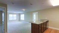 This single-floor space showcases an open-concept layout that seamlessly connects the kitchen and living areas. The kitchen features a prominent island with light granite countertops, an undermount sink with a contemporary faucet, and rich dark-stained cabinetry with brushed metal hardware. The cabinetry provides generous storage, and the granite surface offers ample workspace.

The adjoining living area is expansive, finished with neutral wall-to-wall carpeting that adds warmth underfoot. Soft, neutral-toned walls in a light sage-green hue contribute to a calm and cohesive ambiance throughout. Multiple windows along the far wall allow natural light to filter in, brightening the space and creating an airy, open feel.

Flush-mount ceiling fixtures provide consistent overhead lighting, complementing the natural light from the windows. A doorway visible from the main area leads to an additional room, suggesting versatile functionality. The overall design is clean and understated, with a practical layout suited for everyday living and entertaining.
