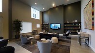 This single-floor space presents a polished, contemporary lounge environment defined by its dramatic ceiling height, which creates an expansive, airy atmosphere. The room is anchored by an olive-toned accent wall hosting a large wall-mounted flat-screen television, flanked on both sides by built-in open shelving that displays carefully curated decorative objects and books, adding a refined, library-like quality to the space.

The seating arrangement is thoughtfully composed, blending a central dark console table with surrounding modular soft seating, including rounded upholstered armchairs in neutral gray tones, cylindrical ottomans, and sleek accent chairs with a modern silhouette. A low-pile area rug in muted tones defines the primary lounge zone, grounding the furniture grouping cohesively.

Natural light enters through a tall, narrow window on the adjacent wall, illuminating a secondary area furnished with a small bistro table and chairs, ideal for individual work or casual meetings. Recessed ceiling lighting supplements the natural light, ensuring consistent, even illumination throughout. The overall ambiance is modern, calm, and professionally designed, suited for both social and focused use.
