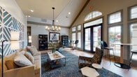 This single-floor leasing office for Landmark Apartments offers a polished, welcoming environment designed to impress visitors from the moment they arrive. The entry foyer sets the tone with warm hardwood-style flooring, soaring vaulted ceilings with exposed beam detailing, and an arched transom window that draws in generous natural light. A striking reception desk anchors the front of the space, finished in teal and white with a bold geometric lattice pattern that is echoed in the decorative ceiling panels above — creating a cohesive, eye-catching focal point.

The lounge area is expansive and thoughtfully furnished, featuring a neutral sectional sofa, woven accent chairs, and a marble-top coffee table arranged atop a richly toned abstract area rug. A statement chandelier with an industrial-modern design hangs overhead, complementing the vaulted ceiling height. One wall showcases a dramatic teal-and-white geometric mural that adds artistic depth to the space.

Large floor-to-ceiling windows and glass-paned French doors line the exterior wall, flooding the interior with natural light and providing views of the outdoor surroundings. A dedicated workstation area with bar-height seating offers a functional spot for consultations. Dark display cabinetry and curated décor accents throughout give the space a refined, residential feel. A private office is accessible from the main floor, completing this well-appointed leasing facility.