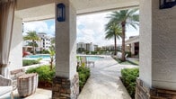 This beautifully designed outdoor amenity space evokes a resort-like atmosphere, seamlessly blending architecture with lush tropical surroundings. The covered lounge area serves as the centerpiece, featuring stucco columns with stone base detailing, warm wood-paneled ceilings, and elegant wall-mounted lantern lighting. Large floor-to-ceiling sliding glass doors connect the covered patio to an interior space, flooding both areas with abundant natural light and creating an effortless indoor-outdoor flow.

The covered patio is appointed with stylish woven lounge chairs, plush cushioned sofas, and wicker accent tables, all set atop travertine tile flooring and anchored by teal-patterned area rugs. Sheer drapery panels frame the seating areas, adding a touch of privacy and sophistication. Lush tropical plantings, including palms and colorful foliage, border the space throughout.

Beyond the covered lounge, a generously sized resort-style pool glistens under open skies, surrounded by additional sun loungers and manicured landscaping. A serene water feature with a fountain is visible in the background, complemented by views of the surrounding residential buildings. The overall ambiance is bright, airy, and inviting, offering a refined outdoor retreat ideal for relaxation and leisure.