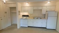 This single-floor, one-bedroom, one-bathroom unit offers a practical and efficient layout designed to maximize usable living space. The kitchen is a central feature, equipped with a white electric coil-burner range, a double-basin sink, and ample white cabinetry providing generous storage above and below the countertops. A range hood above the stove supports ventilation, while the light-colored countertops and white cabinetry contribute to a clean, bright aesthetic. Overhead lighting illuminates the kitchen area with a simple flush-mount ceiling fixture. The flooring transitions from a hard surface in the kitchen to carpeting in the adjacent living area, adding a layer of comfort underfoot. A recessed electrical panel is visible along one wall, and the open sightlines between the kitchen and living space create a connected, airy feel despite the unit's modest scale. The neutral wall tones throughout provide a versatile backdrop suited to a variety of furnishing styles. Overall, this unit offers a no-frills, functional living environment well-suited for those prioritizing efficiency and ease of maintenance.