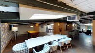 This single-floor space is designed for social gathering and entertainment, blending industrial architectural elements with eclectic, comfortable furnishings. Exposed dark steel beams run across the ceiling alongside visible ductwork and conduit, lending the space an industrial character that is softened by warm pendant lighting and decorative wall sconces. Skylights are positioned throughout, drawing in natural light and brightening the open-plan layout.

The space is organized into several distinct zones. A game area hosts multiple billiard tables illuminated by classic pendant fixtures, providing a dedicated recreation hub. Adjacent to this is a dining and lounge area featuring a large white table surrounded by houndstooth-patterned wicker chairs, ideal for group gatherings. Decorative vases and artwork add curated touches throughout.

A full bar counter with dark granite countertops and upholstered bar stools offers a comfortable spot for refreshments, complete with a microwave and sink. Patterned wallpaper accents add a bold visual element nearby. A separate lounge area centers on a slate-tiled fireplace with an arched opening, flanked by dark upholstered seating and a round ottoman, creating a relaxed and inviting atmosphere. Warm concrete flooring and light wood paneling tie the zones together cohesively.