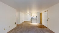 This single-floor residence offers a practical and efficiently designed layout, featuring two bedrooms, two bathrooms, a combined kitchen and living area, and an in-unit laundry room. The open kitchen is well-appointed with white cabinetry accented by warm wood trim, a stainless steel sink, a stainless electric range, a black refrigerator, and a peninsula with counter seating — all set on durable wood-look flooring. A ceiling fan in the adjacent living area enhances comfort, and the space flows naturally between the kitchen and carpeted living zones. The primary bedroom is a comfortable size, featuring plush carpet, a window with natural light, baseboard heating, and a closet with a wood-panel door. The second bedroom is similarly appointed, offering natural light through a window overlooking greenery. Both bathrooms include wood-vanity cabinetry with granite-style countertops, framed mirrors with overhead lighting, and wood-look tile flooring. A dedicated laundry area adds everyday convenience. Throughout the residence, neutral tones, textured ceilings, and consistent finishes create a clean and cohesive aesthetic. A sliding glass door in one of the main rooms opens to an outdoor area, bringing in additional natural light and views of surrounding greenery.