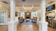 This thoughtfully designed single-floor space combines classic architectural elegance with warm, functional interiors. The centerpiece is a spacious lounge area framed by prominent white decorative columns and a striking coffered ceiling that adds a sense of grandeur and height. A floor-to-ceiling stone fireplace anchors the room, flanked by large windows that flood the space with natural light and offer views of the surrounding greenery. A modern sputnik-style chandelier hangs overhead, complementing the warm-toned wingback chairs and patterned area rug arranged around a central coffee table. Rich hardwood flooring flows throughout the hallway and lounge areas, while a wall-mounted television and a decorative console table provide additional functionality near the entry. The dining room is an intimate retreat featuring a round wooden dining table with upholstered chairs set atop an ornate patterned carpet. Crown molding, a sputnik chandelier, framed artwork, and natural light from large windows complete the refined aesthetic. The private office is a quiet, well-appointed workspace with built-in upper cabinetry, a dedicated desk, patterned carpeting, and ample natural light, making it ideal for focused tasks. Together, these spaces offer a balanced blend of sophistication and practicality.