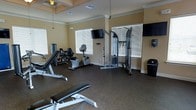 This single-floor fitness center offers a comprehensive workout environment designed to support a range of exercise routines. The space is outfitted with a solid selection of equipment, including a cable resistance machine, a stationary recumbent bike, a leg press machine, and multiple weight benches, providing options for both strength training and low-impact cardio workouts. Two flat-screen televisions are mounted on the walls, allowing members to stay entertained during their sessions. The room is finished with durable rubber flooring, ideal for high-traffic fitness use, and features crown molding and coffered ceiling details that lend a polished, refined look to the space. Warm, neutral-toned walls contribute to a welcoming atmosphere. Multiple large windows fitted with horizontal blinds allow ample natural light to fill the room while maintaining privacy and glare control. Ventilation is supported by ceiling-mounted air vents, ensuring a comfortable environment during workouts. The overall layout is thoughtfully arranged to allow freedom of movement between stations, making it a functional and well-maintained facility suited for everyday fitness use.