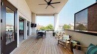 This contemporary single-floor space blends modern interior design with impressive outdoor living. The highlight is a generously sized rooftop deck finished with natural wood planking, offering panoramic views of the surrounding neighborhood. The deck is equipped with a large dining table and chairs, comfortable lounge seating with accent pillows, barrel-style side tables, a wall-mounted flat-screen TV, and large ceiling fans for year-round comfort. Glass and metal railings keep the views unobstructed, while partial overhead coverage provides a sheltered area for gathering. Interior access to the deck is provided through bold navy-blue double glass doors, which also lend the transition areas a clean, architectural character. Inside, the space features high ceilings, light wood plank flooring, and white walls that create a bright, open atmosphere. A dedicated kitchenette area includes warm wood cabinetry, a white countertop, a stainless steel sink with a matte black industrial-style faucet, and a decorative artistic tile backsplash that serves as a visual focal point. The main interior room is open and flexible, highlighted by tall ceilings and glass-paned doors, making it well-suited for events, meetings, or creative uses. The combination of indoor and outdoor areas makes this a versatile and well-appointed space.