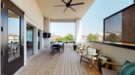 This contemporary single-floor space blends modern interior design with impressive outdoor living. The highlight is a generously sized rooftop deck finished with natural wood planking, offering panoramic views of the surrounding neighborhood. The deck is equipped with a large dining table and chairs, comfortable lounge seating with accent pillows, barrel-style side tables, a wall-mounted flat-screen TV, and large ceiling fans for year-round comfort. Glass and metal railings keep the views unobstructed, while partial overhead coverage provides a sheltered area for gathering. Interior access to the deck is provided through bold navy-blue double glass doors, which also lend the transition areas a clean, architectural character. Inside, the space features high ceilings, light wood plank flooring, and white walls that create a bright, open atmosphere. A dedicated kitchenette area includes warm wood cabinetry, a white countertop, a stainless steel sink with a matte black industrial-style faucet, and a decorative artistic tile backsplash that serves as a visual focal point. The main interior room is open and flexible, highlighted by tall ceilings and glass-paned doors, making it well-suited for events, meetings, or creative uses. The combination of indoor and outdoor areas makes this a versatile and well-appointed space.