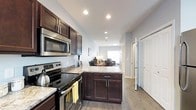This thoughtfully designed multi-floor residence spans three levels, offering a versatile and functional layout suited for everyday living. The kitchen serves as a central highlight, featuring rich dark cabinetry, granite-look countertops, and a full suite of stainless steel appliances including an electric range, built-in microwave, and additional counter space provided by a peninsula that doubles as a prep and serving area. The light gray walls and recessed lighting throughout create a clean, modern ambiance that feels both welcoming and practical. Light wood-tone flooring runs seamlessly from the kitchen into the adjacent living and dining areas, reinforcing an open-concept flow. The living area benefits from natural light streaming through a window at the far end, lending the space a bright and airy quality. A hallway off the kitchen provides access to additional rooms and closets, with crisp white doors adding a fresh contrast to the warm wood tones. Across its three floors, this residence offers ample room for living, entertaining, and private retreat, making it a well-rounded home designed with both style and functionality in mind.