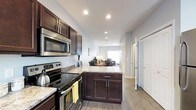 This thoughtfully designed multi-floor residence spans three levels, offering a versatile and functional layout suited for everyday living. The kitchen serves as a central highlight, featuring rich dark cabinetry, granite-look countertops, and a full suite of stainless steel appliances including an electric range, built-in microwave, and additional counter space provided by a peninsula that doubles as a prep and serving area. The light gray walls and recessed lighting throughout create a clean, modern ambiance that feels both welcoming and practical. Light wood-tone flooring runs seamlessly from the kitchen into the adjacent living and dining areas, reinforcing an open-concept flow. The living area benefits from natural light streaming through a window at the far end, lending the space a bright and airy quality. A hallway off the kitchen provides access to additional rooms and closets, with crisp white doors adding a fresh contrast to the warm wood tones. Across its three floors, this residence offers ample room for living, entertaining, and private retreat, making it a well-rounded home designed with both style and functionality in mind.