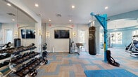 Located at 3240 SW 34th St in Ocala, Florida, this single-floor fitness center is a comprehensively outfitted facility designed to support a wide range of workout styles. The space is divided into distinct functional zones, each thoughtfully arranged and connected by wide corridors with durable wood-look flooring and sky-blue painted walls that lend the entire facility a bright, energetic atmosphere.

The main strength and functional training area features patterned carpet tile flooring in blue and neutral tones, a full dumbbell rack, a heavy punching bag suspended from a bold blue free-standing rig, battle ropes, resistance equipment, a flat bench, and a wall-mounted flat-screen TV flanked by sconce lighting and large mirrors. Arched architectural details and recessed lighting add a polished, upscale character to the space.

An adjoining area hosts a variety of resistance machines, stability balls, and additional free weights, benefiting from abundant natural light streaming through multiple arched windows with outdoor views of the surrounding community.

The dedicated cardio room features wood-look flooring, light blue walls with motivational wall art, a row of treadmills positioned beside windows overlooking the pool area, and ceiling fans to promote airflow. A separate nook accommodates a recumbent bike and additional cardio equipment. Throughout, the design balances functionality with an inviting aesthetic, making this fitness center a standout community amenity.
