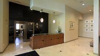 Rosedale Park presents a professionally designed single-floor space anchored by a refined lobby that sets the tone for the entire environment. The reception area is a standout feature, showcasing a rich wood-paneled front desk topped with a glass surface, accented by decorative diamond-shaped hardware details. Overhead, two elegant pendant light fixtures cast a warm, inviting glow throughout the space. A dramatic floor-to-ceiling black accent wall serves as a bold backdrop, displaying the Rosedale Park branding in understated, stylish lettering. The lobby's light-toned tile flooring enhances the sense of openness and cleanliness, while framed artwork adorning the neutral walls adds a cultured, polished touch. A glimpse beyond the reception area reveals additional interior spaces, suggesting a thoughtfully laid-out floor plan. Natural and artificial lighting work in harmony to create a bright, professional ambiance throughout. The overall design balances modern sophistication with a welcoming warmth, making Rosedale Park an inviting destination that conveys both functionality and refined aesthetic sensibility.