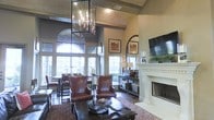 This refined single-floor space makes an immediate impression with its dramatic, soaring ceilings that create an open and airy atmosphere. A striking oversized geometric chandelier serves as a bold focal point, casting warm light across the richly appointed interior. The living area is anchored by a classic white-mantled fireplace, complemented by a wall-mounted flat-screen television and tasteful decorative accents including candles and greenery. Rich leather wingback chairs and a wrought-iron coffee table add a sense of warmth and traditional elegance to the seating arrangement, set atop a patterned area rug over dark hardwood flooring. Expansive arched windows with intricate detailing line the walls, inviting an abundance of natural light and offering views of the lush greenery outside. A large arched mirror adds depth and sophistication to the space. Adjacent to the living area, a dining space with dark wood furnishings continues the cohesive, classic aesthetic. Warm neutral wall tones throughout tie each area together seamlessly. The overall ambiance is one of timeless elegance, combining architectural grandeur with carefully curated furnishings to create a welcoming and visually impressive environment.