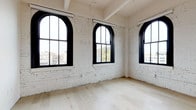 This single-floor loft-style unit masterfully blends industrial heritage with contemporary design. The living areas are defined by whitewashed exposed brick walls and dramatic arched windows with bold black frames, flooding the interior with natural light while offering elevated views of the surrounding cityscape. Light hardwood flooring runs throughout, complementing the bright, airy atmosphere created by the soaring ceilings, which also feature exposed ductwork — a nod to the building's industrial roots. Track lighting provides functional illumination while reinforcing the loft aesthetic.

The kitchen is sleek and modern, outfitted with white flat-front cabinetry, a white subway tile backsplash, light stone countertops, and a full suite of stainless steel appliances including a range, microwave, dishwasher, and refrigerator. A bedroom features a sliding closet door and additional black-framed windows that continue the city views and natural light theme. The bathroom is finished with large-format neutral tile flooring, a white vanity, and a glass-enclosed walk-in shower, offering a clean, minimalist aesthetic. The overall ambiance is bright, open, and urban.
