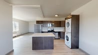 Located on the 13th floor at 707 West 5th Avenue in Spokane, Washington, Unit 1310 is a thoughtfully designed single-floor residence that combines modern finishes with functional living. Light-toned wood-style flooring runs throughout, lending a clean, cohesive feel to the entire space.

The kitchen serves as a central hub of the unit, featuring rich dark-wood cabinetry, stainless steel appliances including a refrigerator, gas range, and over-the-range microwave, and a white quartz-topped island that provides ample prep and dining space. A stacked washer and dryer unit is conveniently integrated into the kitchen area, maximizing utility without sacrificing flow.

The open living area extends naturally from the kitchen, offering generous space bathed in natural light from large windows. The bedroom spaces each feature wide windows with striking elevated views — one overlooking lush treetops and greenery, the other offering sweeping panoramic views of the Spokane cityscape and surrounding skyline, both framed by baseboard heating units beneath each window.

The bathroom is clean and straightforward, with a wall-mounted sink, a mirror with overhead lighting, and a glass-enclosed shower, all finished in neutral tones. The overall ambiance of the unit is bright, modern, and airy, with a practical layout well-suited for comfortable urban living in the heart of Spokane.