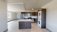 Located on the 13th floor at 707 West 5th Avenue in Spokane, Washington, Unit 1310 is a thoughtfully designed single-floor residence that combines modern finishes with functional living. Light-toned wood-style flooring runs throughout, lending a clean, cohesive feel to the entire space.

The kitchen serves as a central hub of the unit, featuring rich dark-wood cabinetry, stainless steel appliances including a refrigerator, gas range, and over-the-range microwave, and a white quartz-topped island that provides ample prep and dining space. A stacked washer and dryer unit is conveniently integrated into the kitchen area, maximizing utility without sacrificing flow.

The open living area extends naturally from the kitchen, offering generous space bathed in natural light from large windows. The bedroom spaces each feature wide windows with striking elevated views — one overlooking lush treetops and greenery, the other offering sweeping panoramic views of the Spokane cityscape and surrounding skyline, both framed by baseboard heating units beneath each window.

The bathroom is clean and straightforward, with a wall-mounted sink, a mirror with overhead lighting, and a glass-enclosed shower, all finished in neutral tones. The overall ambiance of the unit is bright, modern, and airy, with a practical layout well-suited for comfortable urban living in the heart of Spokane.