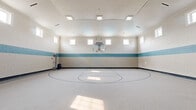 Located at 5475 Winding River Rd in Noblesville, Indiana, this impressive indoor basketball court offers a spacious, single-floor athletic facility designed for serious recreation and sport. The court features a clearly marked playing surface with painted court lines and a center circle, all set atop a durable, speckled rubberized floor that provides both grip and resilience underfoot. A wall-mounted glass backboard and adjustable hoop anchor one end of the court, delivering a professional-grade playing experience. The soaring ceiling height allows for unrestricted play, while a distinctive teal-and-cream painted concrete block wall treatment adds a clean, polished aesthetic throughout the space. Rows of clerestory windows line the upper walls on multiple sides, flooding the interior with abundant natural light and creating an open, airy atmosphere. Recessed and flush-mounted ceiling light fixtures supplement natural light for evening or low-light use. Multiple access doors, including a windowed entry door, provide convenient entry and exit points. A perimeter walkway with a dark speckled floor runs along at least one side of the court, offering additional space for spectators or equipment storage. The overall design is functional, well-maintained, and purpose-built for athletic use.