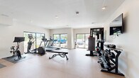 Located at 8895 Perimeter Park Blvd in Jacksonville, FL, this well-appointed fitness center offers a comprehensive workout environment within a efficiently organized single-room layout. The space features bright white walls, recessed lighting, and durable speckled flooring designed to withstand high-traffic exercise use. Large sliding glass windows and floor-to-ceiling panels line multiple walls, flooding the room with natural light and providing open views of the surrounding greenery and landscape outside. The cardio area is equipped with multiple treadmills, an elliptical trainer, and a stationary cycle, all positioned to take advantage of the outdoor views. A multi-functional cable trainer anchors the strength training section, complemented by a fully stocked rack system featuring kettlebells, medicine balls, and resistance accessories — all organized under a bold "Escape Your Limits" frame. A dedicated dumbbell rack offers a wide range of free weights for versatile strength training. An adjustable workout bench provides additional functionality for a variety of exercises. A wall-mounted television and a wall clock add practical amenities to the space. The overall ambiance is clean, modern, and energizing, making this fitness center a well-rounded facility suited for both cardio and strength training routines.