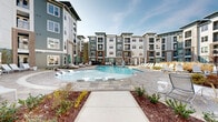 Located at 1515 Rollick Rd in Cary, NC, this contemporary residential community showcases a thoughtfully designed outdoor environment centered around a striking resort-style swimming pool. The expansive pool area is surrounded by rows of lounge chairs and umbrella-shaded seating, creating an inviting retreat for relaxation. The multi-story buildings feature a modern architectural palette of grey and teal board-and-batten siding, brick accents, and white-trimmed windows, with private balconies overlooking the courtyard.

Winding concrete pathways connect the various outdoor amenity zones, lined with carefully curated landscaping including ornamental shrubs, ornamental grasses, and mulched garden beds that provide seasonal color and texture. A dedicated hammock lawn area offers freestanding hammock stands set on lush artificial turf, providing a casual leisure space within the courtyard.

A covered outdoor pavilion serves as a social hub, featuring a herringbone-patterned stone patio, comfortable lounge seating, and dining tables with chairs — ideal for gatherings or everyday outdoor living. Additional seating areas and pergola structures are distributed throughout the grounds, enhancing the community's walkable, amenity-rich character. The cohesive design and well-maintained grounds reflect a high standard of community living in the heart of Cary, North Carolina.