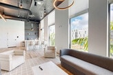 Located at 20775 NW 17th Ave in Miami Gardens, Florida, this contemporary lounge space delivers a striking blend of industrial and modern design across a single, open floor. Soaring ceilings with exposed ductwork and conduit lend an urban, loft-inspired character, while a dramatic circular pendant light anchors the room with warm, ambient illumination. Sleek pendant lights hang above the wet bar and kitchen area, adding focused task lighting throughout.

The living area is generously appointed with a curated mix of seating — including a grey curved sofa, plush cream armchairs, and rust-toned upholstered ottomans — all arranged around a central coffee table atop a textured area rug. A large flat-screen television is mounted against a sophisticated textured accent wall in muted slate tones, creating a stylish focal point.

The wet bar and kitchenette area features white cabinetry, open shelving with decorative accents, and a clean white island with bar seating — ideal for casual gatherings. Light wide-plank hardwood flooring runs throughout, brightening the space and complementing the neutral palette.

Floor-to-ceiling windows along the exterior wall flood the interior with abundant natural light and frame lush tropical palm views and the surrounding Miami Gardens landscape. Black-framed glass entry doors complete the modern aesthetic, enhancing the sense of openness and connectivity.