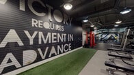This expansive fitness facility is thoughtfully designed to accommodate a wide range of training styles within a single, open-plan floor. The space makes an immediate impression with its bold, large-scale motivational mural — "Victory Requires Payment in Advance" — displayed prominently on a dark feature wall alongside an artificial turf lane, ideal for functional training and sled drills.

The strength training zone is generously outfitted with cable machines, plate-loaded equipment, adjustable benches, free weights, barbells, and squat racks, including striking red power racks that serve as visual anchors throughout the space. A dedicated cardio area lines one side of the facility, featuring treadmills, ellipticals, and stationary bikes positioned beside large windows that invite natural light and outdoor views through exposed brick columns.

The functional training section includes medicine balls, battle ropes, a heavy tire, and rowing machines, supported by floor-to-ceiling mirrors that enhance both spatial depth and training feedback. Rubber flooring throughout ensures durability and safety across all zones.

The industrial aesthetic is reinforced by an exposed ceiling with visible ductwork and pendant lighting, creating a raw yet polished atmosphere. The layout is open and navigable, offering ample space between equipment and a cohesive flow throughout the entire facility.