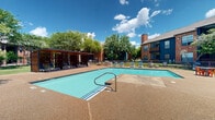 Located at 6000 Ohio Dr in Plano, Texas, this outdoor amenity space is a well-appointed community gathering area designed for relaxation and recreation. At its centerpiece is a generously sized swimming pool with clear turquoise water, accessible via a classic handrail entry and surrounded by a textured concrete pool deck. The deck provides ample space for lounge chairs, allowing residents to enjoy the Texas sunshine in comfort.

Adjacent to the pool is a modern slatted wood pergola structure that offers shaded seating, creating a cool retreat on warm days. A covered pavilion with a pitched roof sits nearby, providing additional shelter and serving as a gathering point for social activities. Outdoor seating and lounging areas are arranged throughout the patio, complementing the open-air atmosphere.

The surrounding grounds are lushly landscaped with well-maintained green lawns and mature deciduous trees that provide generous shade and a sense of natural privacy. The multi-story brick residential buildings frame the space, featuring classic brick exteriors with pitched rooflines that lend the community a timeless, traditional aesthetic. The combination of manicured greenery, inviting water features, and thoughtfully designed shade structures creates a vibrant and welcoming outdoor environment ideal for both leisure and community gatherings.
