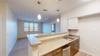 This single-floor unit offers a thoughtfully designed, open layout that integrates the kitchen and living areas seamlessly. The kitchen is equipped with light wood cabinetry, stainless steel appliances including a microwave and range, and a wraparound peninsula with neutral countertops that doubles as a casual dining or workspace area. Pendant lighting hangs above the peninsula, complementing the track lighting that illuminates the open living space. Multiple windows allow natural light to filter throughout, offering views of surrounding greenery.

The bedrooms are carpeted and well-proportioned, each featuring windows with blinds that bring in ample daylight and outdoor views of trees. One bedroom includes access to a walk-in closet, providing generous storage. The bathrooms are finished with stone-look tile flooring, dark wood vanity cabinetry, and white countertops. One bathroom features a walk-in shower with grab bars, while another includes additional accessibility-focused grab bars near the toilet, indicating the unit's suitability for a range of mobility needs. The overall design is clean, neutral, and functional throughout.