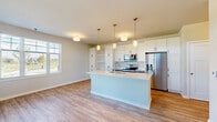 Located at 1774 W Orchard Rd in North Aurora, Illinois, this single-floor residence combines modern design with functional living spaces. The open-concept kitchen serves as the heart of the home, showcasing white shaker-style cabinetry, a large center island with a light quartz countertop, and stainless steel appliances including a French door refrigerator with ice dispenser, electric range, built-in microwave, and dishwasher. Three pendant lights with brass accents illuminate the island, complementing the warm-toned hardwood-style flooring that flows throughout the main living areas. The living space is bathed in natural light through large windows and a glass door that offer views of the surrounding landscape. Bedrooms feature plush neutral carpeting and generous closet space, including a walk-in closet with built-in shelving and hanging rods. A dedicated utility or laundry area provides tall white cabinetry for ample storage. The bathroom is finished with a gray shaker-style vanity, white quartz countertop, a large mirror, and contemporary globe lighting. A well-lit hallway connects the kitchen to the bedrooms and additional rooms, maintaining a cohesive, clean aesthetic with neutral tones and white trim throughout the entire home.