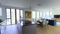 This single-floor space showcases a thoughtfully designed open-plan layout that seamlessly integrates the kitchen and dining areas into one cohesive living environment. Large floor-to-ceiling windows wrap around the space, flooding the interior with abundant natural light and offering expansive views of the surrounding urban landscape.

The kitchen is anchored by a substantial central island featuring dark granite countertops that contrast elegantly with the white cabinetry below. A modern stainless steel faucet adds a sleek, functional touch, while pendant lighting descends from the white ceiling above, enhancing both form and function. The clean lines of the cabinetry reflect a contemporary minimalist aesthetic throughout.

Transitioning naturally from the kitchen, the dining area accommodates a generously sized rectangular table surrounded by white upholstered chairs, making it well-suited for both everyday dining and entertaining. A framed abstract artwork on the wall adds a tasteful decorative accent between the two zones.

Light hardwood flooring runs continuously throughout, unifying the space and complementing the bright, airy atmosphere created by the expansive glazing. The overall ambiance is modern, open, and inviting.
