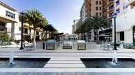 This exceptional outdoor amenity space delivers a resort-like experience designed for relaxation and entertainment. At its heart lies a large, pristine swimming pool with a striking water feature that serves as the focal point of the entire area. A central island platform with stepping stones bridges across the pool, creating a dramatic architectural element that connects the space visually and physically.

Flanking the pool are two fully equipped outdoor kitchen stations, each outfitted with professional-grade stainless steel grills and generous counter space, ideal for outdoor cooking and entertaining. Surrounding these stations, comfortable seating areas with dining tables and chairs invite residents to gather and socialize in an open-air setting.

Tall, mature palm trees are thoughtfully positioned throughout the space, providing a tropical ambiance and natural shade. A covered pergola-style lounge area is visible to one side, offering a shaded retreat with additional seating. The entire deck is finished in light-toned pavers that complement the warm, sun-drenched atmosphere.

The space is framed by elegant, multi-story buildings featuring balconies and Mediterranean-inspired architecture, enhancing the upscale character of the environment. With abundant natural light, lush landscaping, and high-end amenities, this outdoor space offers a sophisticated and inviting atmosphere throughout the day.