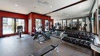 This single-floor fitness center is designed to support a comprehensive workout experience in a well-appointed, energizing environment. The space is defined by its bold red accent walls, which contrast sharply against crisp white ceilings and dark-trimmed architectural details, creating a dynamic and motivating atmosphere throughout.

The main exercise area is generously proportioned, featuring durable rubber flooring suited for high-traffic fitness use. A wide array of cardio equipment is arranged throughout the room, including multiple treadmills, elliptical trainers, and recumbent and upright cycles. A dedicated free weights section includes a full rack of dumbbells paired with a flat bench, while floor space accommodates additional functional training. Full-length mirrors along one wall visually expand the space and support proper form during workouts.

Natural light fills the room through large, dark-framed windows and glass doors that offer views of an outdoor pool area, enhancing the open and airy feel of the space. Ceiling fans and recessed lighting provide comfortable illumination at all hours.

A separate anteroom adjacent to the main floor includes a water cooler for hydration and convenient access to a dedicated restroom, making the facility fully self-contained. The overall design balances aesthetic appeal with practical functionality, offering users a polished and complete fitness environment.