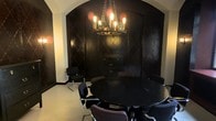 This single-floor space makes a bold design statement through its striking use of dark, dramatic aesthetics. The focal point of the room is a rounded black table surrounded by sleek black chairs with chrome frames, creating a sophisticated yet edgy gathering space ideal for meetings or intimate dining. An ornate wrought-iron chandelier with exposed Edison bulbs hangs from above on a chain, casting a warm, ambient glow that contrasts beautifully against the deep, dark surroundings. The walls are finished in near-black tones and feature a textured diamond-pattern detail, adding depth and visual interest to the space. A built-in dark wood cabinet or credenza is seamlessly integrated into the background, providing both storage and a refined architectural element. The arched alcove framing the seating area is rendered in a lighter plaster or stucco finish, offering a compelling contrast to the dark interior and lending a vaulted, cave-like character to the room. Light-toned flooring grounds the space and prevents the overall scheme from feeling overwhelming. The result is a cohesive, intentionally designed environment with a rich, moody ambiance.