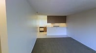 This single-floor space features a straightforward, functional layout designed to maximize usability. The living area is finished with wall-to-wall carpeting in a neutral tone, creating a comfortable and cohesive atmosphere. A textured popcorn ceiling runs throughout, adding a classic residential character to the space.

The kitchen is separated from the living area by a half-wall peninsula that doubles as a breakfast bar, offering a convenient pass-through that connects the two spaces while maintaining a sense of openness. The kitchen itself is finished with light tile flooring, providing a practical and easy-to-maintain surface. Cabinetry in warm wood tones lines the kitchen, complemented by under-cabinet appliances and a sink positioned near a window fitted with vertical blinds, allowing natural light to filter in.

A dark overhead soffit detail above the kitchen peninsula adds a distinctive architectural touch, framing the transition between the living and kitchen zones. The overall color palette is neutral and clean, featuring light walls and trim throughout. A ceiling vent ensures climate control across the living space. The layout and design make this a practical, move-in-ready space suited for everyday living.