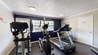 This fitness center is organized across two connected rooms, offering a well-rounded workout environment. The cardio area features bold navy accent walls, large multi-pane windows that bring in natural light and outdoor views, and dark carpet flooring. Equipment includes multiple treadmills and elliptical machines arranged to maximize the available space. Motivational wall decals, including phrases like "Everyday Is a Good Day" and "Work Harder," contribute to an energizing atmosphere. The textured ceiling with crown molding and flush-mount lighting provides a clean, finished look throughout.

The adjoining strength training room transitions to lighter gray walls and continues the dark flooring. It houses a Life Fitness cable machine, a leg press/hack squat apparatus, and a weight bench, offering options for resistance-based workouts. A large wall mirror enhances the sense of space and supports form monitoring during exercise. A wall-mounted television is also present for entertainment during workouts. Both rooms are accessible via standard interior and exterior doors, and the overall layout supports a comfortable, functional fitness experience.