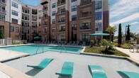 Located at 4451 Vilana Rd in Raleigh, NC, this outdoor amenity space is thoughtfully designed to offer a resort-style living experience within a modern residential community. The centerpiece of the space is a generously sized swimming pool with a smooth tile surround, lined with lounge chairs and shade sails, creating an ideal setting for relaxation under the open sky. Surrounding multi-story buildings with private balconies frame the courtyard, contributing to a sense of enclosure and community.

The expansive courtyard is paved with decorative stone pavers in a natural stone pattern, providing a durable and visually appealing surface throughout. Multiple seating areas are arranged across the patio, including teal-upholstered lounge chairs, a sectional sofa with accent pillows, and metal dining tables with chairs — all arranged for both intimate gatherings and larger social occasions.

A built-in outdoor grilling station is conveniently positioned near the seating areas, making the space well-suited for outdoor entertaining. Tall evergreen trees are planted throughout the courtyard, adding vertical greenery and a sense of privacy. A gated entry leads directly to the pool area, and a covered porte-cochère with decorative planters marks the main building entrance, completing this well-appointed outdoor retreat.