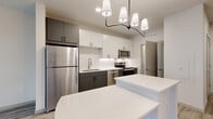 This well-designed one-bedroom, one-bathroom unit offers a thoughtful layout across a single floor, making efficient use of its compact footprint. The open-concept kitchen serves as the centerpiece of the living area, featuring two-tone cabinetry with dark lower cabinets and white uppers, a white subway tile backsplash, quartz countertops, and a full suite of stainless steel appliances including a refrigerator, dishwasher, range, and microwave. A white kitchen island with generous counter space anchors the room and is illuminated by a contemporary multi-pendant black chandelier. Light wood-style plank flooring flows seamlessly throughout the unit, contributing to a clean, modern aesthetic. The bedroom is a comfortable, well-proportioned space with a large window fitted with roller shades, offering views of an interior courtyard and pool area. A dedicated closet with built-in wire shelving provides ample storage. The unit also includes a conveniently located in-unit laundry closet with a stacked washer and dryer. The bathroom features matching gray cabinetry with matte black hardware and a white quartz countertop, consistent with the home's cohesive modern design. Neutral white walls and abundant recessed lighting complete the bright and functional interior.