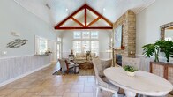 This thoughtfully designed single-floor space exudes a relaxed coastal aesthetic throughout its well-appointed layout. The architectural centerpiece is the main living area, where a dramatic vaulted ceiling with exposed rich-toned wood truss beams draws the eye upward, complemented by a white-painted tongue-and-groove ceiling that amplifies the sense of height and openness. A floor-to-ceiling stacked stone fireplace anchors one wall, providing a striking focal point and a warm, natural contrast to the soft sage-green and pale blue-grey wall tones found throughout the space.

Large multi-pane windows flood the interior with abundant natural light, offering views of the surrounding outdoors while sheer white curtains add an airy, breezy quality. The living room flows seamlessly into the dining area, both finished with neutral stone tile flooring that reinforces the coastal palette. Beadboard wainscoting lines several walls, adding classic architectural detail and texture.

A separate dining/lounge room with teal-toned walls provides a more intimate gathering space, complete with built-in bench seating and wall-mounted television. The kitchen features granite countertops, warm wood cabinetry, a mosaic tile backsplash, and ample counter space. A hallway connects the various areas, maintaining cohesive flow throughout this welcoming, light-filled space.