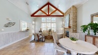This thoughtfully designed single-floor space exudes a relaxed coastal aesthetic throughout its well-appointed layout. The architectural centerpiece is the main living area, where a dramatic vaulted ceiling with exposed rich-toned wood truss beams draws the eye upward, complemented by a white-painted tongue-and-groove ceiling that amplifies the sense of height and openness. A floor-to-ceiling stacked stone fireplace anchors one wall, providing a striking focal point and a warm, natural contrast to the soft sage-green and pale blue-grey wall tones found throughout the space.

Large multi-pane windows flood the interior with abundant natural light, offering views of the surrounding outdoors while sheer white curtains add an airy, breezy quality. The living room flows seamlessly into the dining area, both finished with neutral stone tile flooring that reinforces the coastal palette. Beadboard wainscoting lines several walls, adding classic architectural detail and texture.

A separate dining/lounge room with teal-toned walls provides a more intimate gathering space, complete with built-in bench seating and wall-mounted television. The kitchen features granite countertops, warm wood cabinetry, a mosaic tile backsplash, and ample counter space. A hallway connects the various areas, maintaining cohesive flow throughout this welcoming, light-filled space.