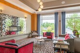 This sophisticated single-floor amenity space is thoughtfully designed to serve as a premier social and recreation hub. The open layout is anchored by distinct activity zones that flow seamlessly into one another across hardwood-style flooring and richly patterned area rugs. A full-size billiards table occupies a central position, while a dedicated shuffleboard table stretches along one side of the space beneath tasteful wall sconces and textured column accents.

The lounge area commands attention with a linear fireplace framed by a bold, metallic mixed-media art installation, accented by warm gold wall panels and a cascading globe pendant light fixture. Seating includes a mix of gray and crimson upholstered sofas and armchairs arranged around marble-top coffee tables, creating an inviting gathering atmosphere.

A built-in wet bar, clad in warm wood tones and stacked-stone tile detailing, features a mirrored backsplash, under-counter refrigeration, a sink, and a mounted flat-screen television overhead. Adjacent to the bar, a bistro-style café table near floor-to-ceiling windows offers views of mature trees and landscaped surroundings, allowing generous natural light to fill the space. Tray ceilings with crown molding lend architectural depth throughout, completing the polished, upscale character of the space.