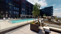 This expansive outdoor amenity space is defined by its contemporary industrial-inspired architecture, combining dark metal cladding, light brick masonry, and oversized steel-framed windows that create a bold, urban aesthetic. The centerpiece is a generously sized, resort-style swimming pool with crystal-clear turquoise water, bordered by sleek glass panel railings that preserve sightlines across the deck. A wide pool surround finished in large-format stone-look tile provides ample space for rows of lounge chairs and shaded umbrella seating, ideal for relaxation and recreation.

Adjacent to the pool deck, a separate elevated outdoor terrace extends along the building's upper level, featuring wide-plank wood-toned decking that adds warmth against the structure's dark facade. Floor-to-ceiling glass doors and expansive windows connect this terrace seamlessly to an interior lounge space, allowing natural light to flow freely between indoors and out. The terrace offers sweeping panoramic views of the surrounding cityscape and open sky, enhancing the sense of openness and elevation.

Curated landscaping, including mature trees set in large dark metal planters and low ground-cover plantings, introduce organic texture throughout the space. Comfortable seating arrangements, including lounge sofas and woven accent chairs, are thoughtfully positioned to encourage both social gatherings and quiet retreat, completing this well-appointed outdoor environment.