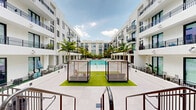 Located at 24 NE First Ave in Dania Beach, Florida, this outdoor amenity level is designed to deliver a resort-style experience within a modern multi-story residential community. The centerpiece of the space is a generously sized swimming pool with crystal-clear turquoise water, framed by wide white porcelain tile decking and lined with lounge chairs ideal for sunbathing. Tall, mature palm trees are strategically placed throughout the courtyard, providing natural shade and reinforcing the tropical South Florida aesthetic. Large raised planter beds with lush tropical foliage serve as elegant landscaping dividers throughout the pool deck. A dedicated sun lounge area features shaded cabana structures set over manicured artificial turf, offering a comfortable retreat from the sun. The surrounding multi-story building displays a clean, contemporary white façade with black-railed balconies and floor-to-ceiling glass doors at the ground level, creating a seamless visual connection between interior and exterior spaces. Wooden-treaded staircases with sleek dark metal railings provide access between levels, blending warmth with modern design. The overall ambiance is bright, open, and vacation-inspired, making this amenity level a defining feature of the Dania Beach property.
