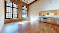 This single-floor loft unit showcases a thoughtful industrial conversion, preserving the architectural integrity of its original structure while incorporating contemporary finishes throughout. The main living area commands immediate attention with its dramatically tall ceilings, exposed brick walls, and large arched multi-pane windows that flood the space with abundant natural light. Original hardwood flooring with rich, warm tones runs throughout, complementing the raw, industrial aesthetic defined by an exposed ductwork system and structural timber beams overhead.

The kitchen is well-appointed with light maple cabinetry, speckled granite countertops, and a full suite of stainless steel appliances including a range, over-the-range microwave, dishwasher, and refrigerator. An upper mezzanine-style loft area is accessible and adds an additional layer of vertical interest to the space.

Private rooms feature neutral grey walls and the same consistent hardwood flooring, offering a calm contrast to the open living areas. The bathroom presents light tile flooring, a vanity with clean white countertops, a bathtub with shower, and a distinctive rustic timber post—a nod to the building's historic origins. Built-in shelving and a dedicated electrical panel further enhance the unit's functional design.