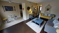 This single-floor space showcases an open-concept layout that seamlessly connects the living area to the kitchen. The living room features rich, dark hardwood flooring anchored by a light gray area rug, creating a warm contrast against the neutral beige walls. A rustic pallet-style coffee table serves as a focal point, complemented by a mustard yellow tufted sofa adorned with decorative pillows. An industrial-style console table with a decorative lamp and ornate mirror adds character to the wall, while additional decorative mirrors and wall accents contribute to the eclectic aesthetic. The transition from the living area to the kitchen is marked by a shift to light tile flooring, where dark cabinetry and a countertop are visible. A small bistro-style table and stool near the kitchen entrance offer a casual dining option. A hallway visible from the living room leads to additional areas of the unit. The space benefits from recessed lighting and natural light filtering in from windows at the far end, creating a bright and inviting atmosphere throughout. The overall design blends industrial, rustic, and vintage elements for a distinctive, personalized ambiance.