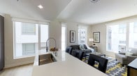 This contemporary single-floor space showcases a thoughtfully designed open-concept layout that effortlessly blends the kitchen and living areas into one cohesive, functional environment. The kitchen features a sleek white quartz countertop island with a stainless steel undermount sink and a professional-style spring-neck faucet, offering both style and practicality. Clean white cabinetry and modern finishes complete the culinary space. The living area flows naturally from the kitchen, anchored by a comfortable dark sofa, black leather accent chairs, and a patterned area rug in navy and yellow tones that adds a vibrant focal point to the room. Framed artwork adorns the walls, contributing to the curated, modern aesthetic. A warm table lamp provides ambient lighting alongside the generous natural light that streams in through multiple large windows dressed with white roller shades. The neutral color palette of whites and soft grays throughout creates a clean, airy atmosphere, while the open sightlines between rooms enhance the overall sense of spaciousness. The space is well-suited for both relaxed everyday living and casual entertaining.