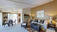 This single-floor residence offers a thoughtfully designed layout centered around a comfortable and stylishly appointed living area. The main living space features warm earth-toned walls that create an inviting ambiance, complemented by plush carpeting underfoot and a decorative area rug with rich, bold patterns. The room is furnished with a neutral sofa accented by patterned throw pillows, a wicker accent chair with cushioned seating, and a dark wooden chest that serves as a functional coffee table centerpiece. Wall art adds a refined, curated touch to the space. Natural light filters in from a sliding glass door that opens to an outdoor balcony, where additional seating is visible, extending the living area outdoors. A ceiling fan overhead provides both practical ventilation and a casual aesthetic detail. Transitioning from the living area, the adjacent kitchen is accessible through an open passageway, featuring cabinetry and standard appliances in a compact, efficient layout. The overall design of the space balances comfort and style, with warm lighting, cohesive color tones, and carefully selected furnishings that create a welcoming and livable environment throughout.