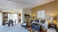 This single-floor residence offers a thoughtfully designed layout centered around a comfortable and stylishly appointed living area. The main living space features warm earth-toned walls that create an inviting ambiance, complemented by plush carpeting underfoot and a decorative area rug with rich, bold patterns. The room is furnished with a neutral sofa accented by patterned throw pillows, a wicker accent chair with cushioned seating, and a dark wooden chest that serves as a functional coffee table centerpiece. Wall art adds a refined, curated touch to the space. Natural light filters in from a sliding glass door that opens to an outdoor balcony, where additional seating is visible, extending the living area outdoors. A ceiling fan overhead provides both practical ventilation and a casual aesthetic detail. Transitioning from the living area, the adjacent kitchen is accessible through an open passageway, featuring cabinetry and standard appliances in a compact, efficient layout. The overall design of the space balances comfort and style, with warm lighting, cohesive color tones, and carefully selected furnishings that create a welcoming and livable environment throughout.