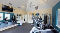 Located in Lewisville, this single-floor fitness center is thoughtfully designed to support a well-rounded workout experience. The space features vaulted ceilings that lend an open, airy feel, complemented by a ceiling fan that promotes comfortable air circulation during exercise sessions. The light-toned walls and recessed lighting create a bright, welcoming environment conducive to focused training. The gym is equipped with a selection of cardio machines, including ellipticals and stationary bikes, positioned to maximize use of the floor space. A cable-based strength training machine provides options for resistance exercises, while a dedicated free weights and bench area supports strength conditioning. Along one wall, a well-organized rack displays an assortment of medicine balls in various weights, catering to functional and dynamic training routines. Full-length mirrors line one side of the room, allowing users to monitor their form throughout their workouts. The durable dark flooring is designed for high-traffic athletic use, providing both safety and ease of maintenance. A set of glass-paneled doors offers a visual connection to adjacent areas, adding to the overall sense of openness. This fitness center is a practical, well-equipped facility suited for a variety of fitness goals and training styles.