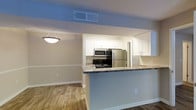 This single-floor residence showcases a thoughtfully designed open-concept layout that seamlessly connects the kitchen and living areas. The kitchen is a standout feature, equipped with stainless steel appliances including a refrigerator, microwave, and range. Crisp white cabinetry provides ample storage, while the dark granite-style countertops offer a striking contrast that adds depth and elegance to the space. A peninsula counter extends into the living area, creating a natural divide between the two spaces while also serving as a casual dining or prep area. Warm hardwood flooring runs throughout, lending a cohesive and inviting feel to the interior. A decorative pendant light hangs above the dining area, adding a touch of character to the otherwise clean and modern aesthetic. The walls are finished in a soft neutral tone, complemented by white trim and baseboards that frame the space neatly. Ceiling vents are strategically placed for efficient climate control. The overall ambiance is bright, clean, and functional, making this a comfortable and practical living space suited for everyday living.