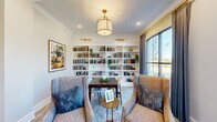 Located at 1670 Friendship Rd in Hoschton, Georgia, this single-floor combined dining and living room offers a well-appointed and intimate setting designed for both relaxation and conversation. The room's most prominent feature is a floor-to-ceiling built-in white bookcase that spans the entirety of one wall, fitted with brass picture lights that highlight curated books, decorative objects, and potted greenery. Crown molding trims the ceiling throughout, lending a refined architectural detail that enhances the room's polished aesthetic. A brass drum pendant light anchors the space from above, complemented by recessed lighting that ensures even, warm illumination throughout. The seating arrangement features upholstered wingback chairs in neutral tones accented with teal throw pillows, arranged around a dark wood dining table and a small round brass accent table. Soft, patterned area rugs define and unify the living and dining zones. Large black-framed windows dressed with layered drapes allow generous natural light to fill the room while providing expansive views of the surrounding landscaped grounds and treeline. An open arched doorway connects the space to an adjacent corridor, contributing to an open and flowing layout. The overall ambiance is elegant yet approachable.